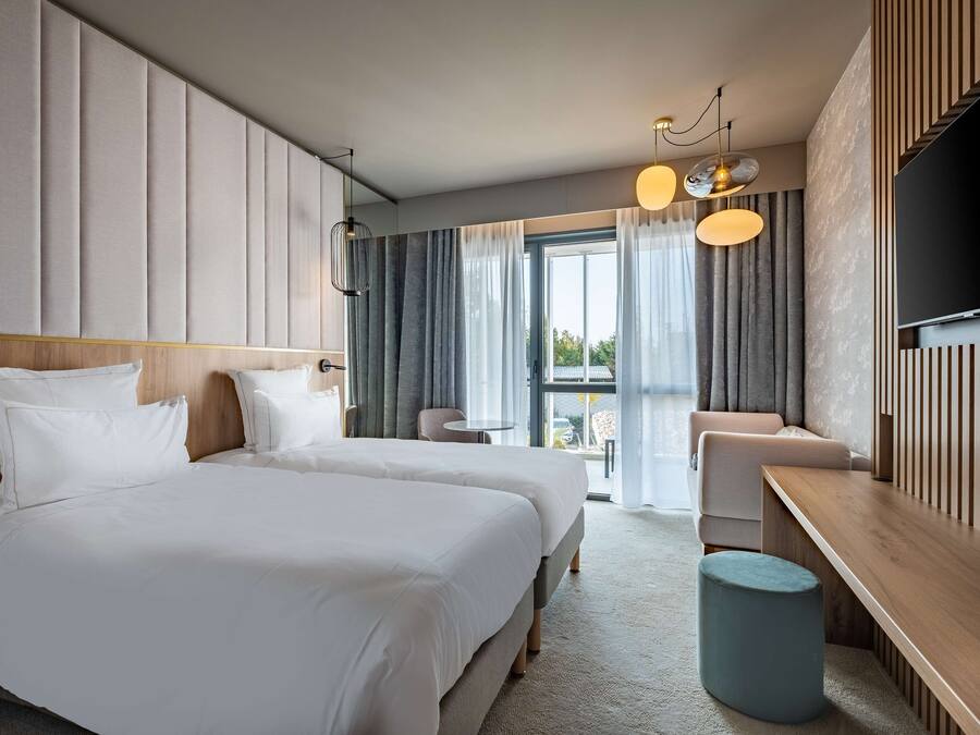 A modern hotel room with two beds, a wooden bench, a round table, and a TV mounted on the wall.