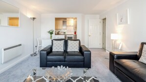 A modern living room with a black leather sofa, a glass coffee table, and a kitchen in the background. - Roomspace Apartments - Regents Court (Kingston Upon Thames)