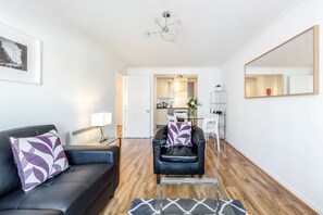 A modern living room with a black leather sofa, a glass coffee table, and a dining area with a white chair and a table. - Roomspace Apartments - Regents Court (Kingston Upon Thames)