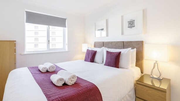 A neatly made bed with white and red bedding, a bedside lamp, and a window with blinds. - Roomspace Apartments - Regents Court (Kingston Upon Thames)