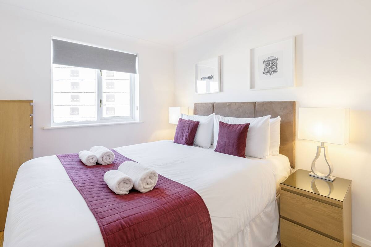 A neatly made bed with white and red bedding, a bedside lamp, and a window with blinds.