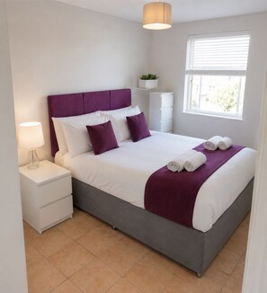 A neatly made bed with white and purple bedding, a bedside lamp, and a window with blinds.