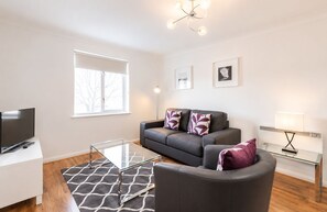 A modern living room with a sofa, a glass coffee table, a television, and a lamp. - Roomspace Apartments - Regents Court (Kingston Upon Thames)