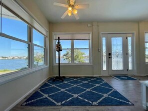A spacious room with large windows offering a view of the water, a ceiling fan, and a blue patterned rug.