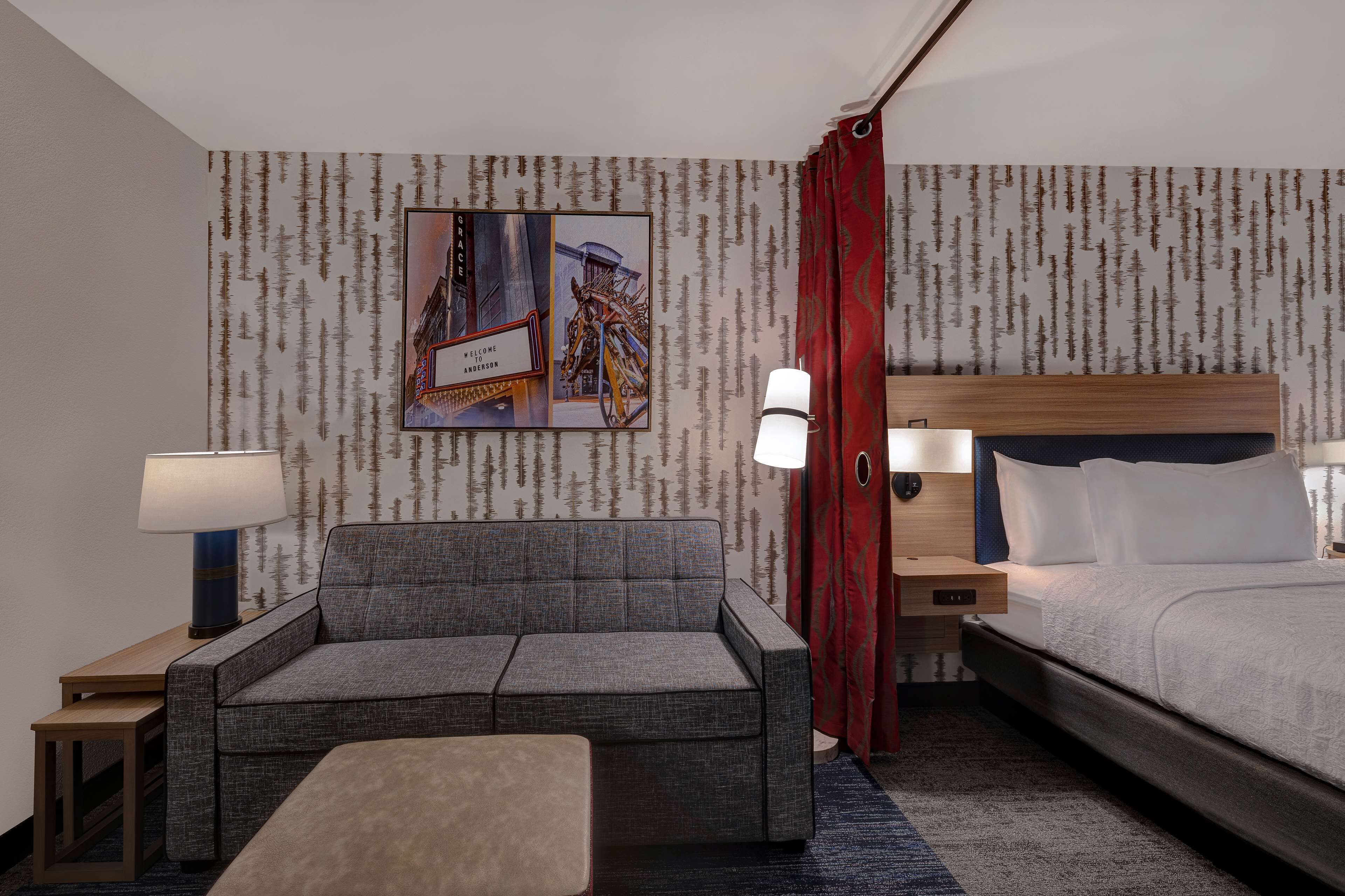 A hotel room with a grey sofa, a bed, a wooden nightstand, a lamp, and a wall with a patterned wallpaper.
