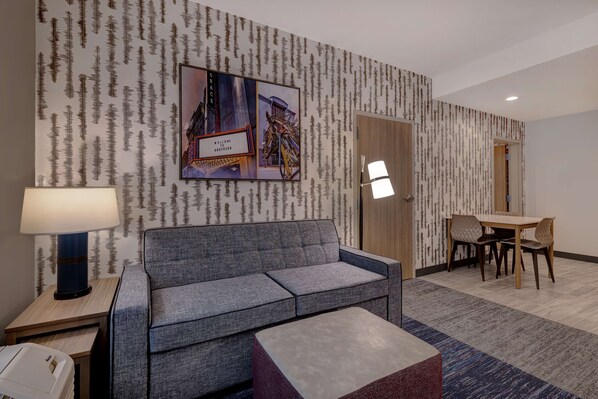 A hotel room with a grey sofa, a small table with a lamp, a dining area with a table and chairs, and a wall with a forest pattern and a welcome sign.