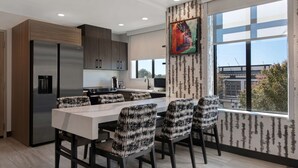 A modern kitchen with a large island, dark cabinets, and a view of the city through large windows. - Home2 Suites by Hilton Anderson Downtown (Anderson)