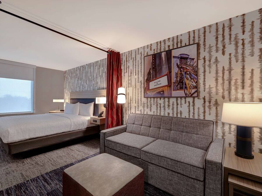 A hotel room with a bed, a sofa, a small table, a nightstand with a lamp, and a wall with a forest wallpaper.