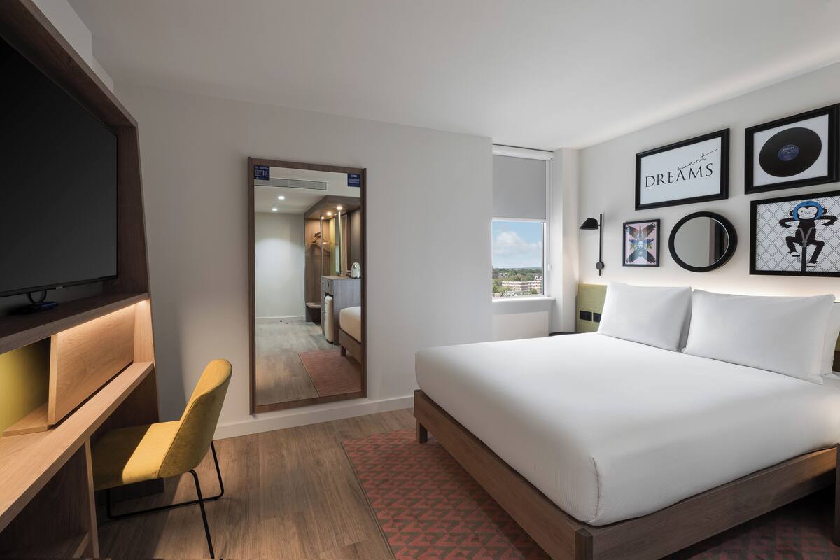 A modern hotel room with a large bed, a desk with a chair, a TV, and artwork on the walls.