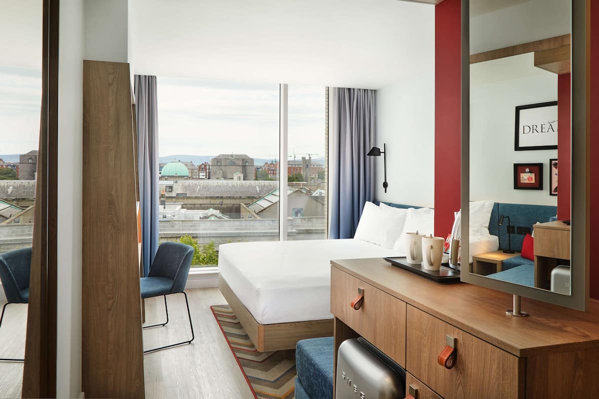 A modern hotel room with a large bed, a desk with a computer, a chair, and a view of the cityscape.