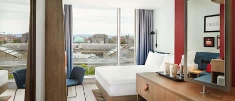 A modern hotel room with a large bed, a desk with a computer, a chair, and a view of the cityscape.