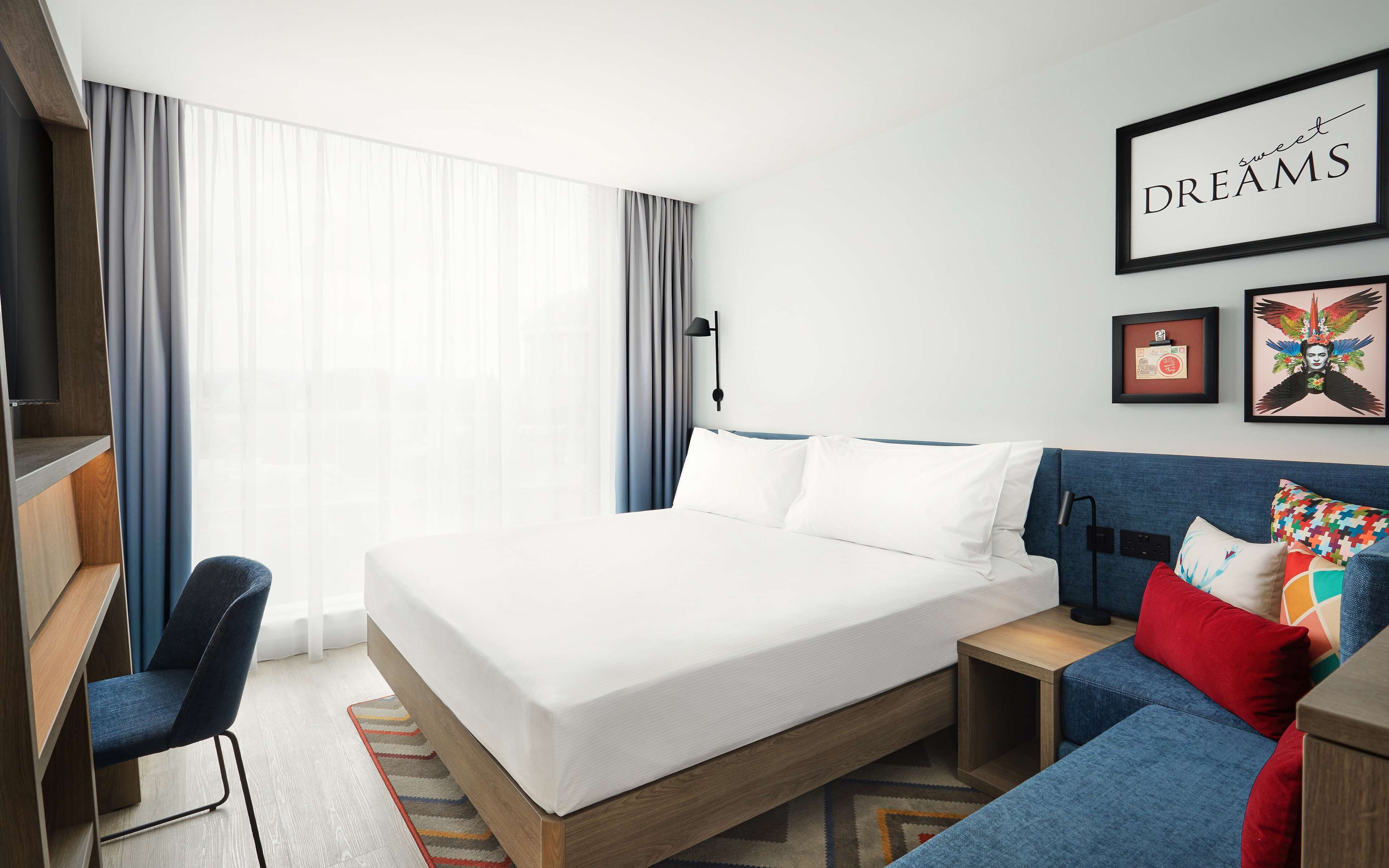 Una habitación de hotel moderna con una cama grande, un escritorio, una silla, un sofá y obras de arte en las paredes.