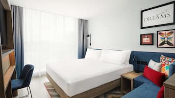 Una habitación de hotel moderna con una cama grande, un escritorio, una silla, un sofá y obras de arte en las paredes.