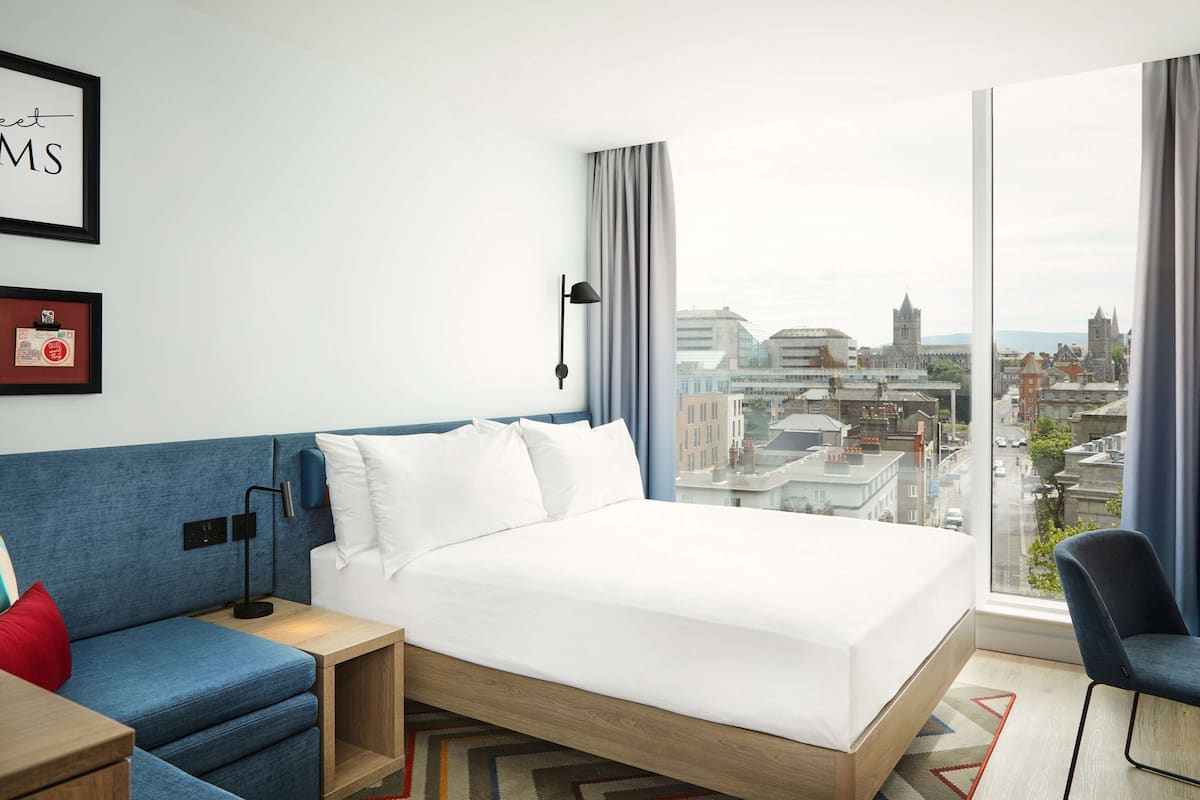 A hotel room with a large bed, a blue sofa, a desk, and a chair. There is a city view through the window.