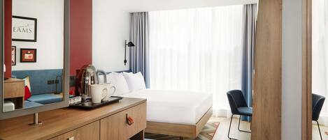 A hotel room with a bed, a desk with a coffee maker, a chair, and a large window with curtains.
