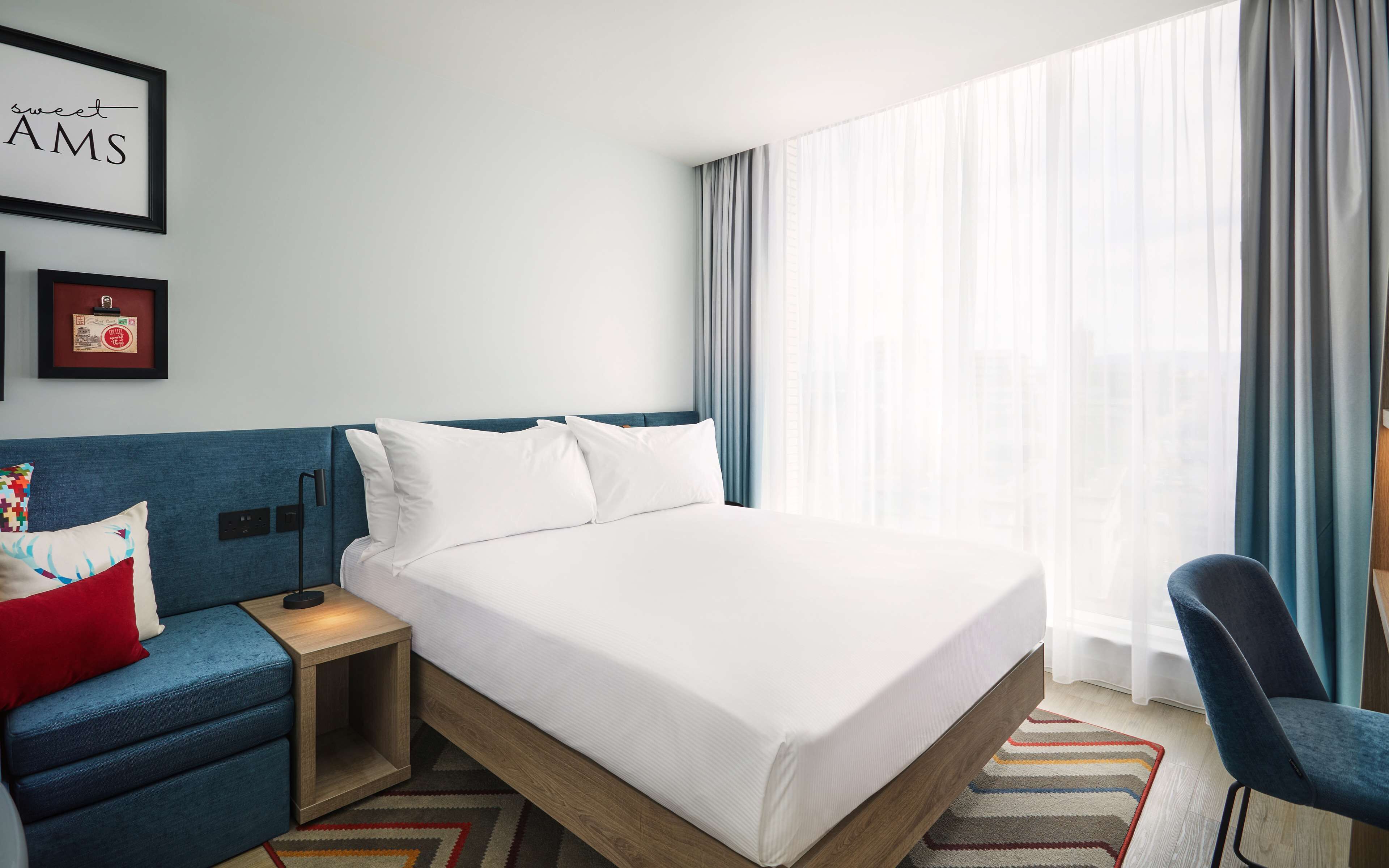 A hotel room with a bed, a nightstand, a chair, a blue sofa, and a window with curtains.