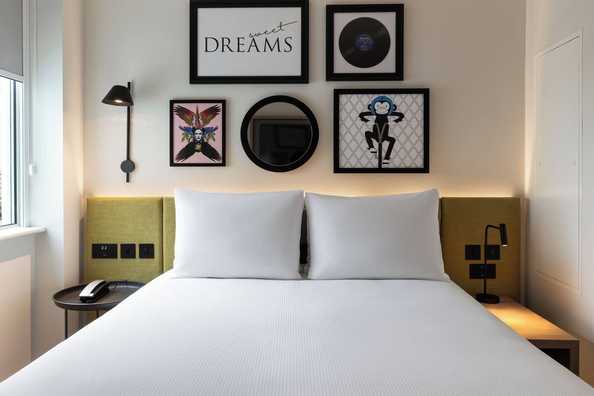 A hotel room with a large bed, a bedside table with a lamp, a phone, and framed artwork on the wall.