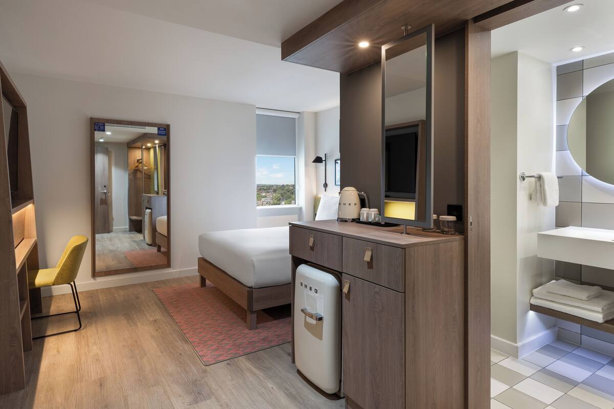 A modern hotel room with a bed, a small refrigerator, a desk with a chair, a large mirror, and a bathroom with a sink and towel rack.