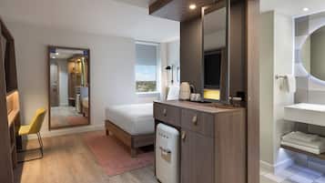 Una habitación de hotel moderna con una cama, un refrigerador pequeño, un escritorio con una silla, un espejo grande y un baño con lavamanos y perchero para toallas.