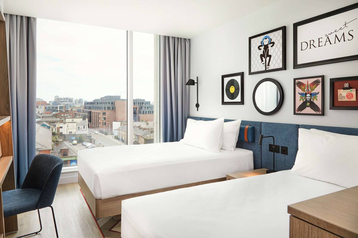 A hotel room with two beds, a desk, and a chair. There are framed pictures on the wall, including one with the word "DREAMS" and another with a monkey.