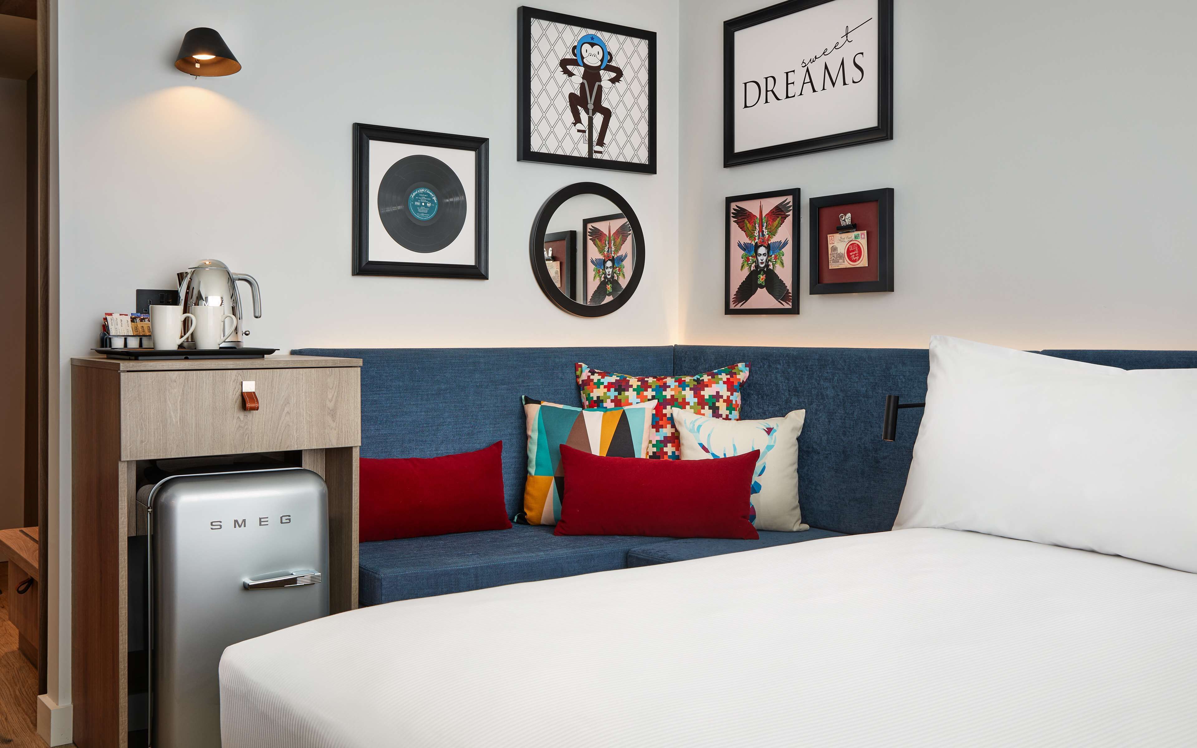 Quarto de hotel moderno com uma cama, uma pequena cozinha e uma parede decorada com obras de arte emolduradas.