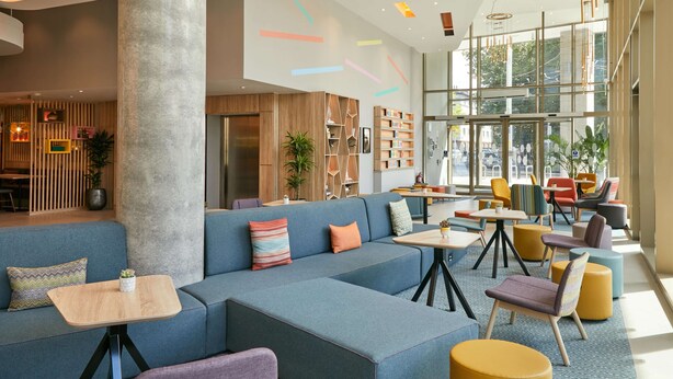 A modern lounge area with a large blue sofa, small tables, colorful chairs, and a large window providing a view of the city. - Hampton by Hilton Dublin City Centre (Dublin)