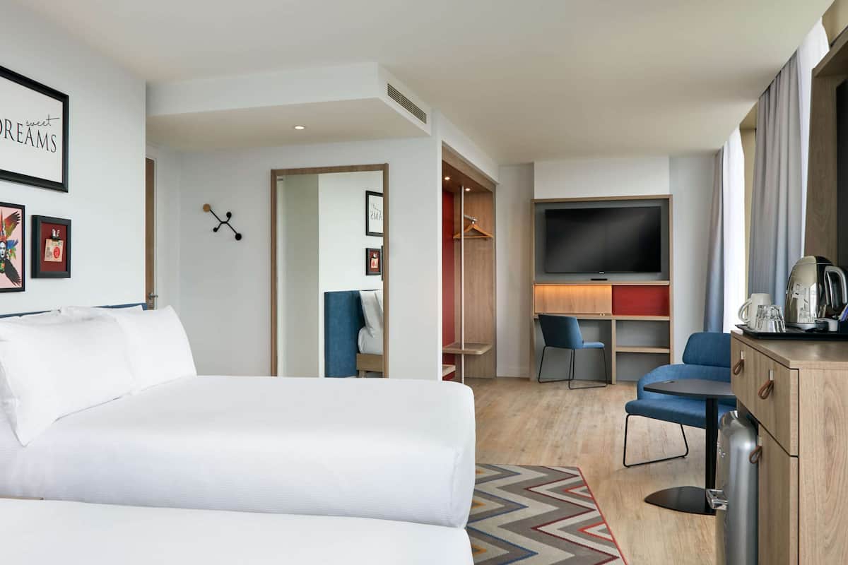 A modern hotel room with a large bed, a desk with a chair, a TV, and a coffee machine.