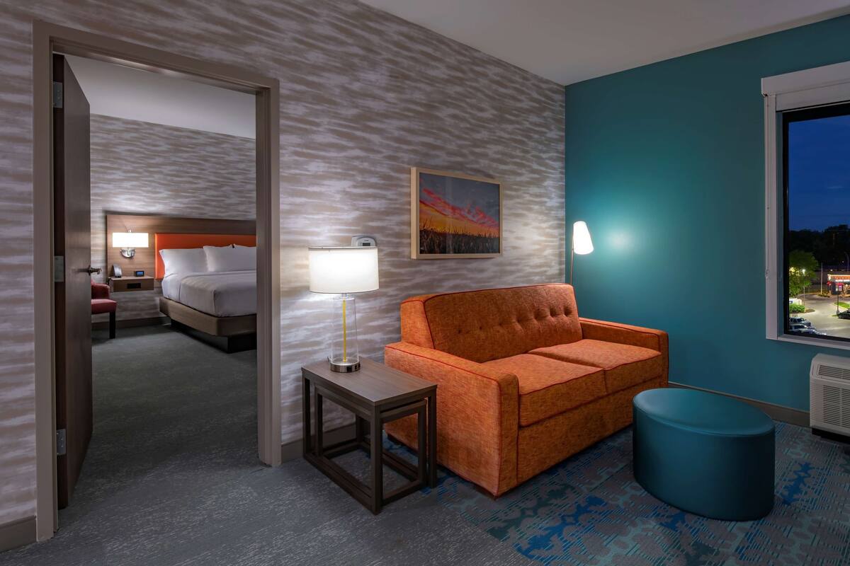 A hotel room with a bed, a sofa, a small table, a lamp, a painting, and a window with a view of the city at night.