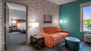A hotel room with a bed, a sofa, a small table, a lamp, a painting, and a window with a view. - Home2 Suites by Hilton Omaha I-80 at 72nd Street, NE (Omaha)