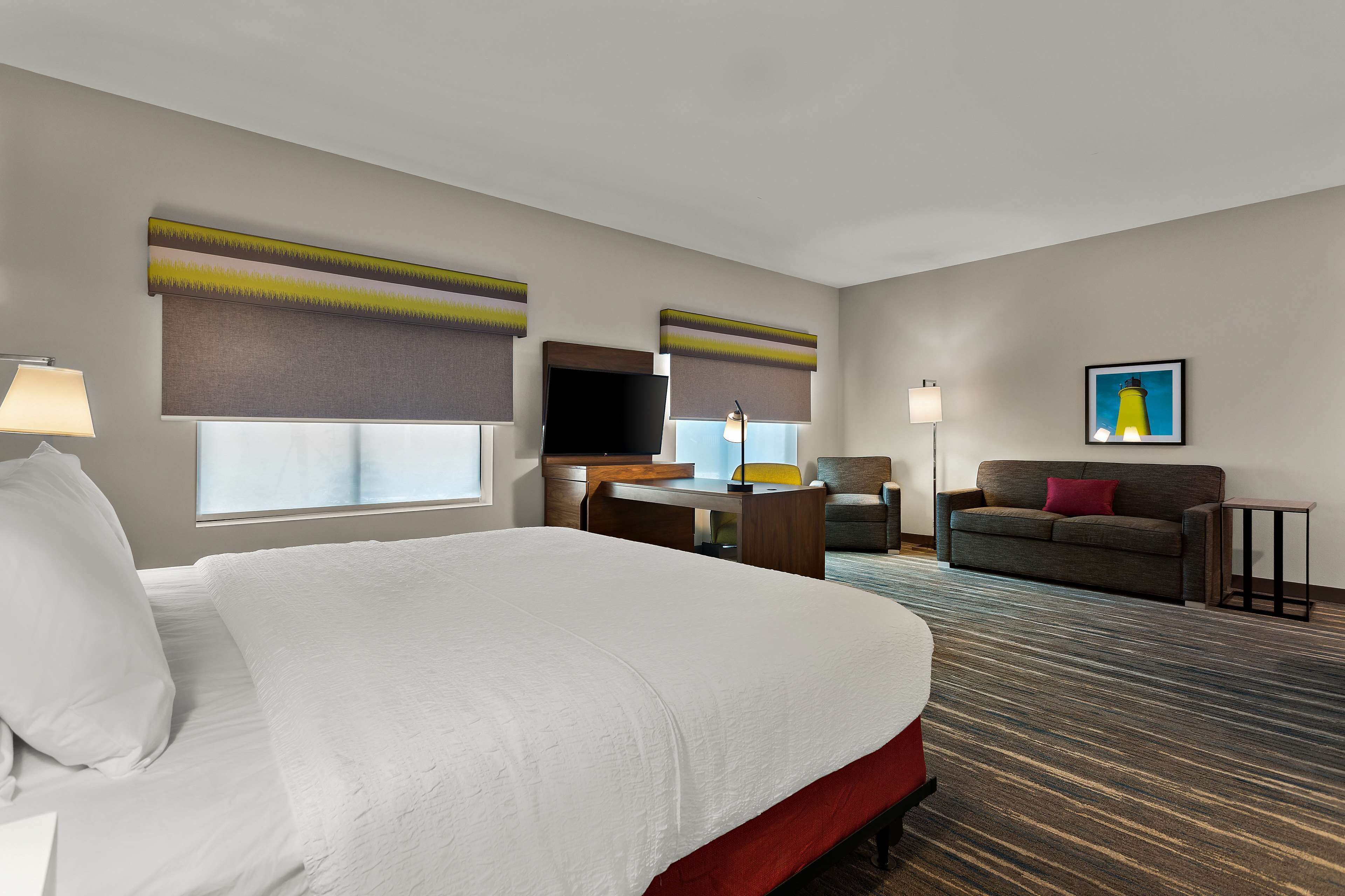 Quarto de hotel com uma cama grande, uma escrivaninha com cadeira, um sofá, uma televisão e obras de arte nas paredes.