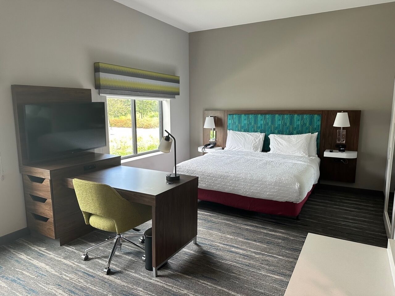 A hotel room with a bed, desk, chair, TV, bedside lamps, and a window.