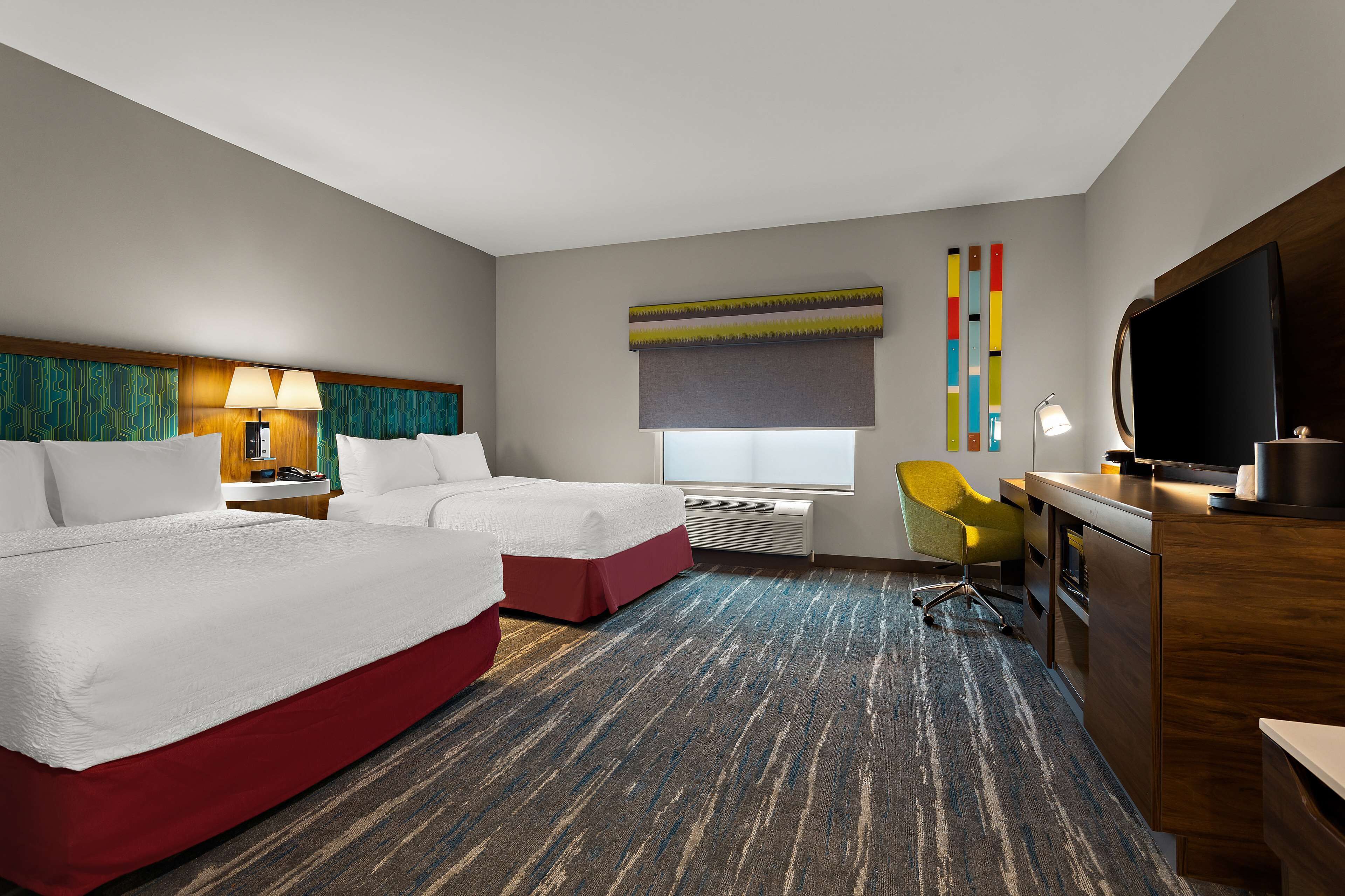 A hotel room with two beds, a desk, a chair, a TV, and a window with blinds.