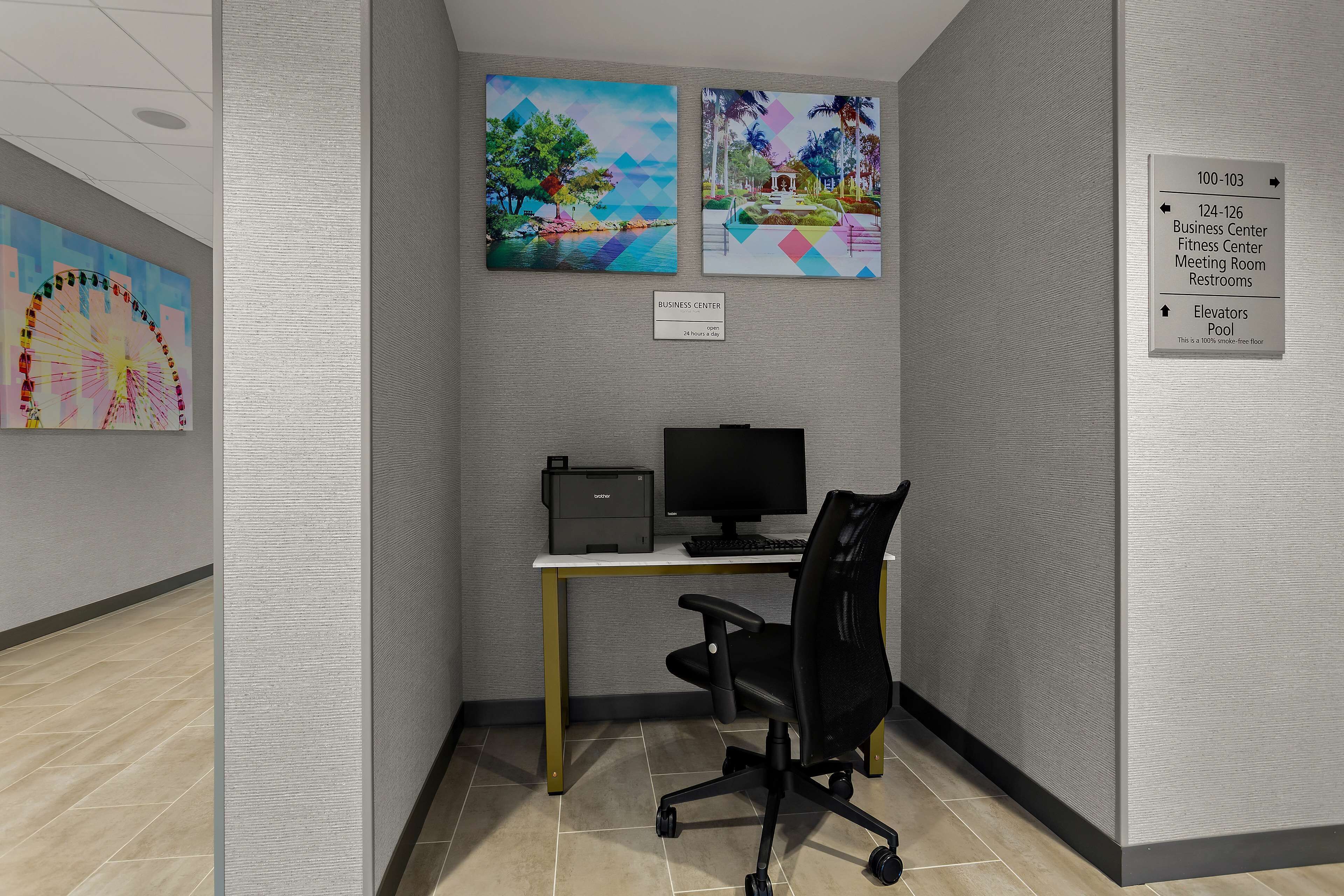 A business center with a desk, computer, and office chair. There are two paintings on the wall and a sign indicating the location of the business center, elevators, and pool.
