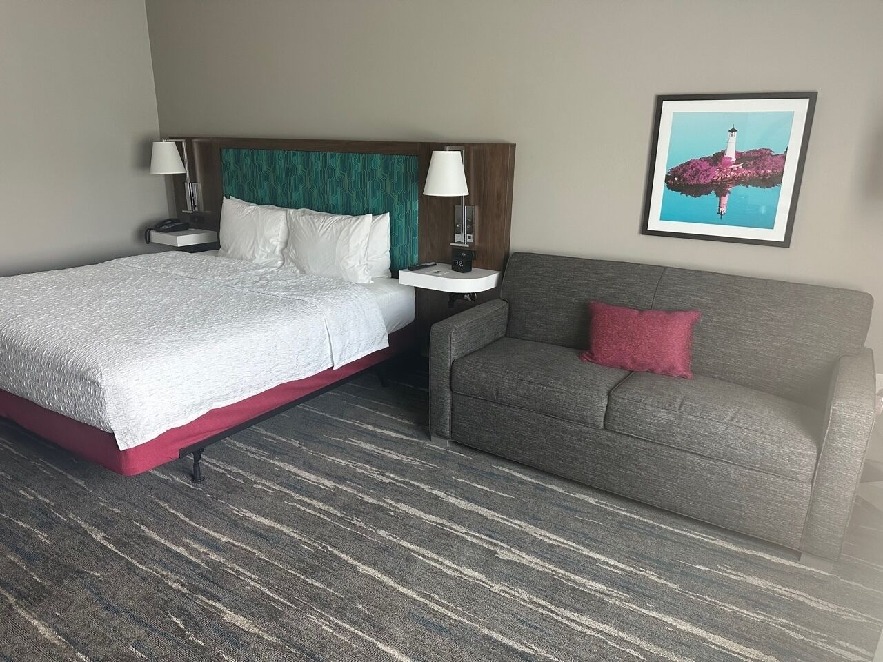 A hotel room with a bed, a grey sofa, a teal headboard, a framed picture on the wall, and a patterned carpet.