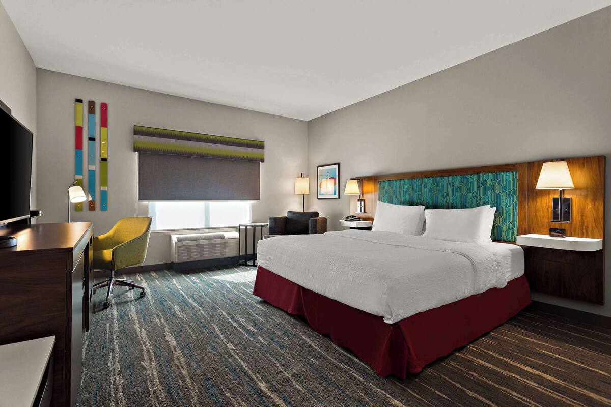 A hotel room with a large bed, a desk, a chair, a TV, and a window with blinds.
