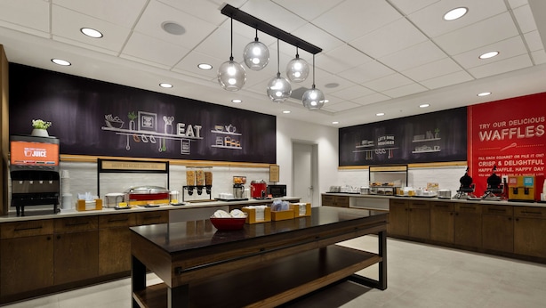 A modern cafe with a central wooden table, a juice dispenser, and a menu board on the wall. - Hampton Inn & Suites Ruskin I-75 (Ruskin)