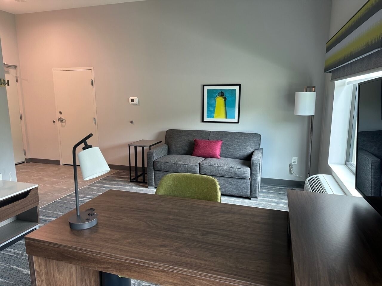 Une chambre d’hôtel moderne avec un bureau en bois, un canapé gris, un fauteuil vert, une œuvre d’art encadrée représentant un phare et une fenêtre donnant sur un paysage.