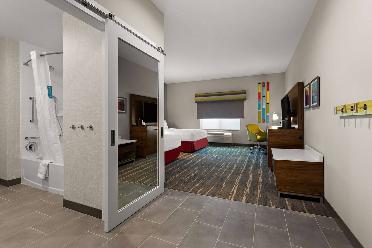 A hotel room with a sliding door leading to a bathroom, a bed with a red accent, a desk with a chair, and a large mirror.