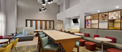 A modern hotel lobby with a large wooden table, multiple seating areas, and a large wall with a screen.