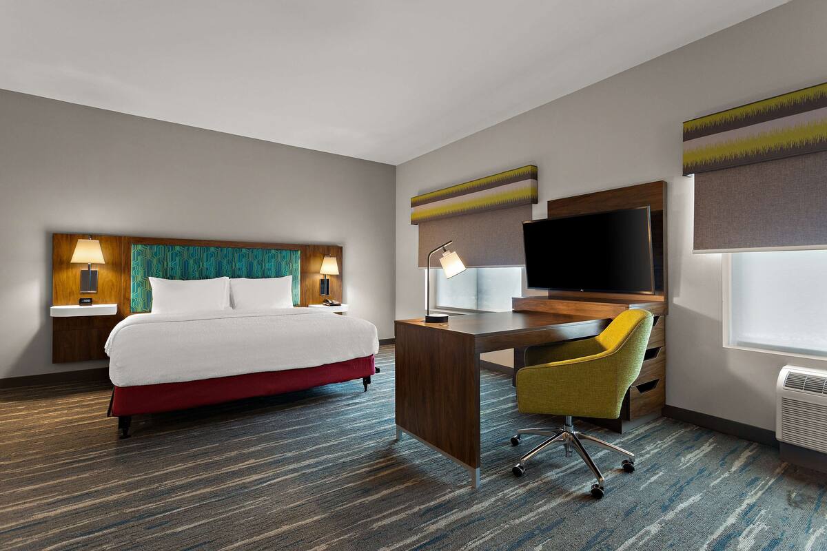 A hotel room with a bed, desk, chair, TV, and wall art.