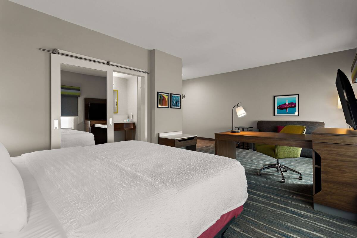 A hotel room with a large bed, a desk, a chair, and a sofa. There are two framed pictures on the wall and a lamp on the desk.