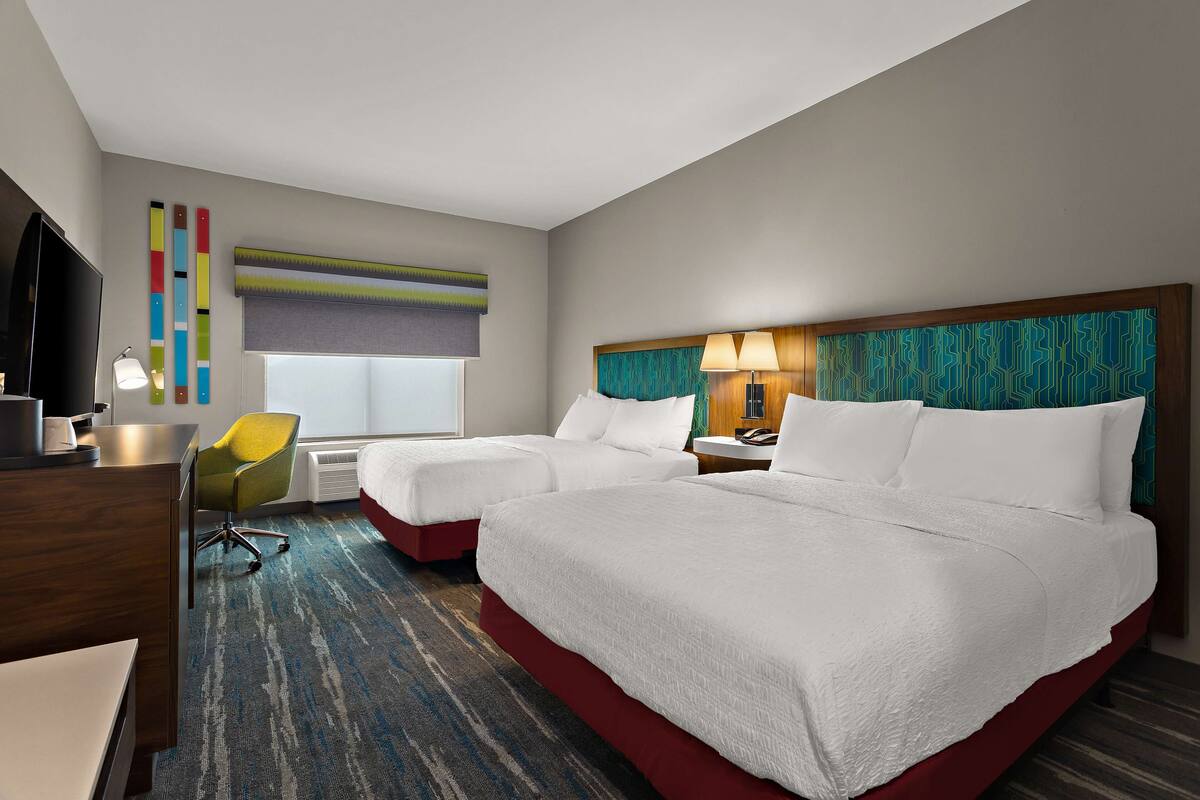 A hotel room with two beds, a desk, a chair, a TV, and a window with blinds.