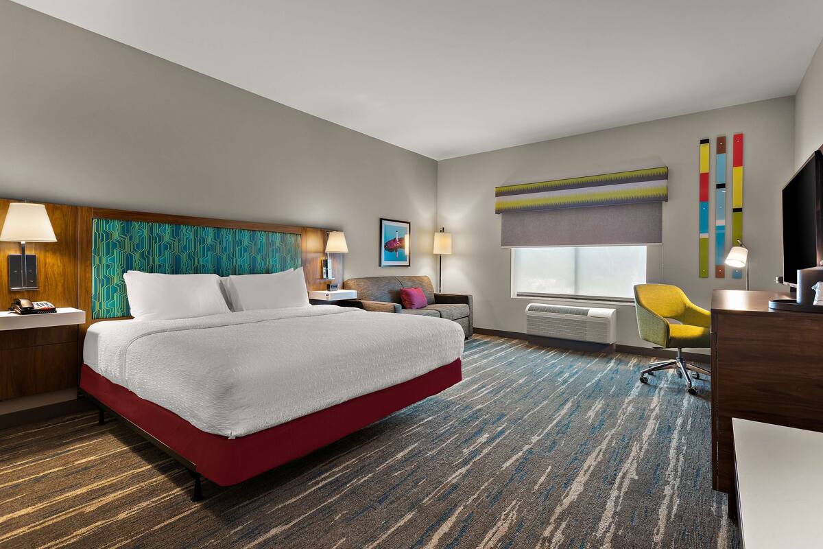 A hotel room with a large bed, a desk, a chair, a window with blinds, and a colorful wall decoration.