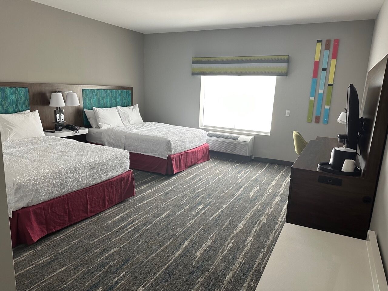 A hotel room with two beds, a desk, a chair, a TV, and a window with blinds.