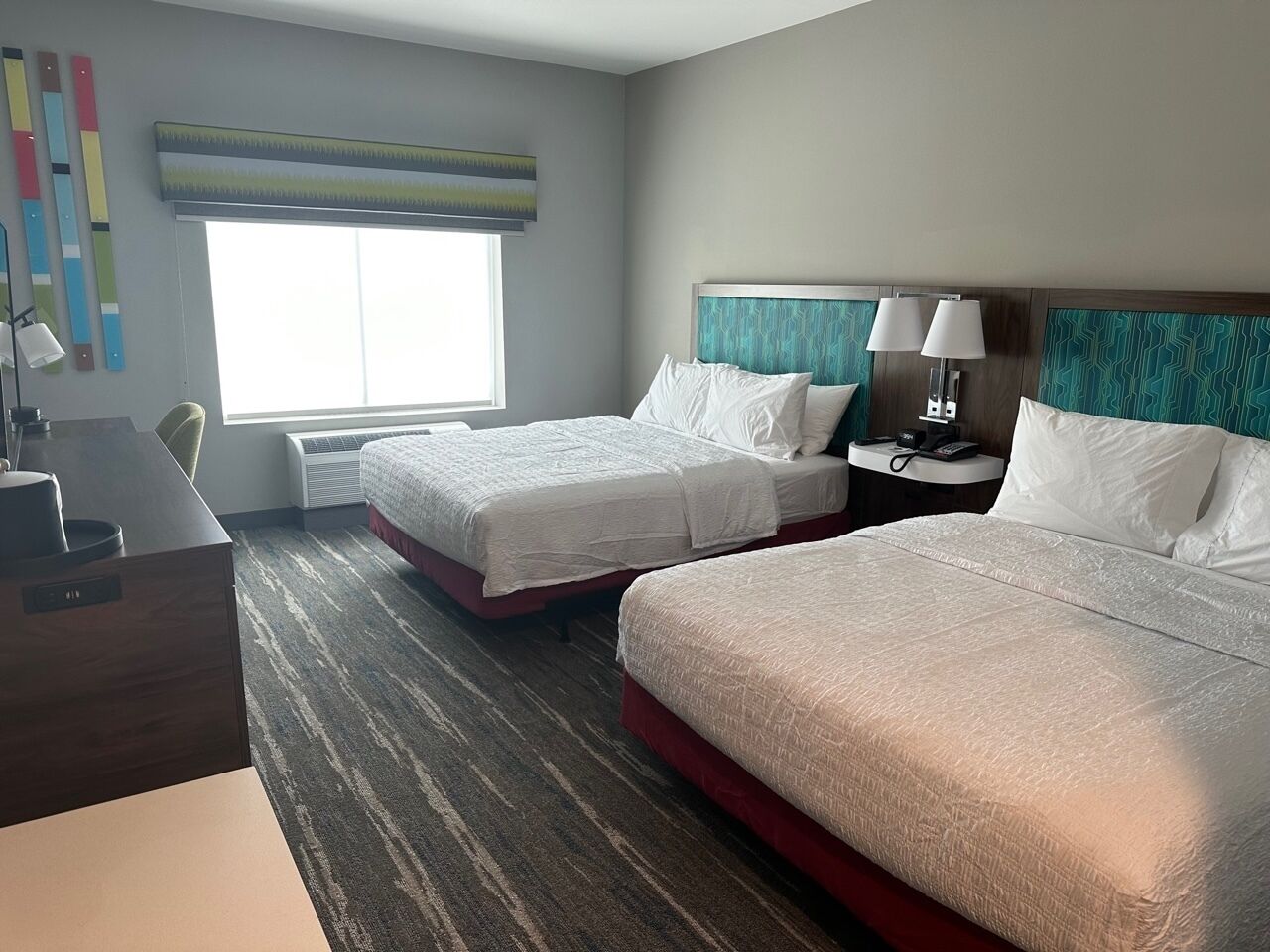 Habitación de hotel con dos camas, un escritorio, una silla, una lámpara y un ventanal con persianas.