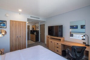 Una habitación de hotel con una cama, un escritorio con silla, un televisor y una kitchenette al fondo.