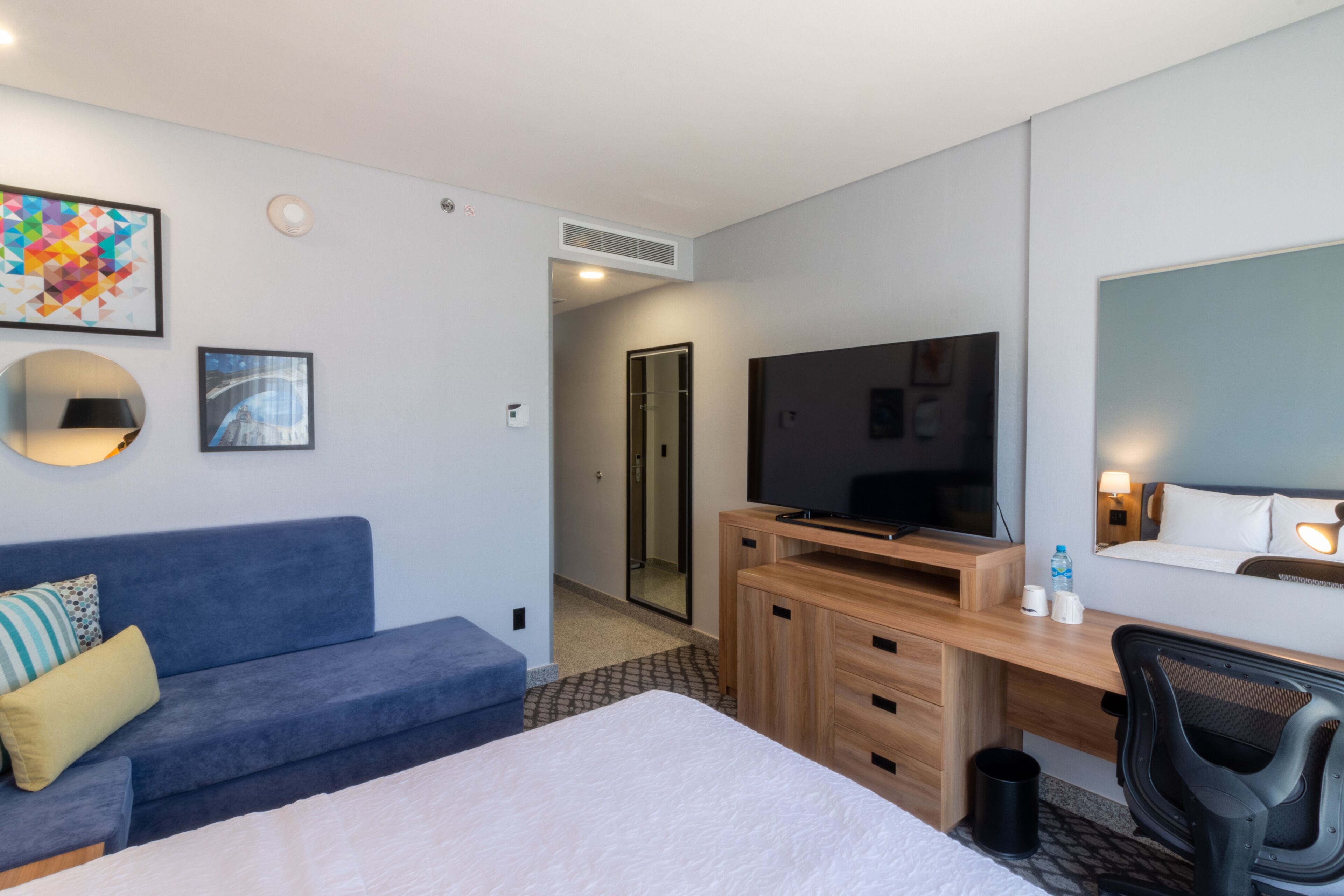 Ein modernes Hotelzimmer mit einem großen Bett, einem blauen Sofa, einem Holztisch mit Fernseher und einem Spiegel.