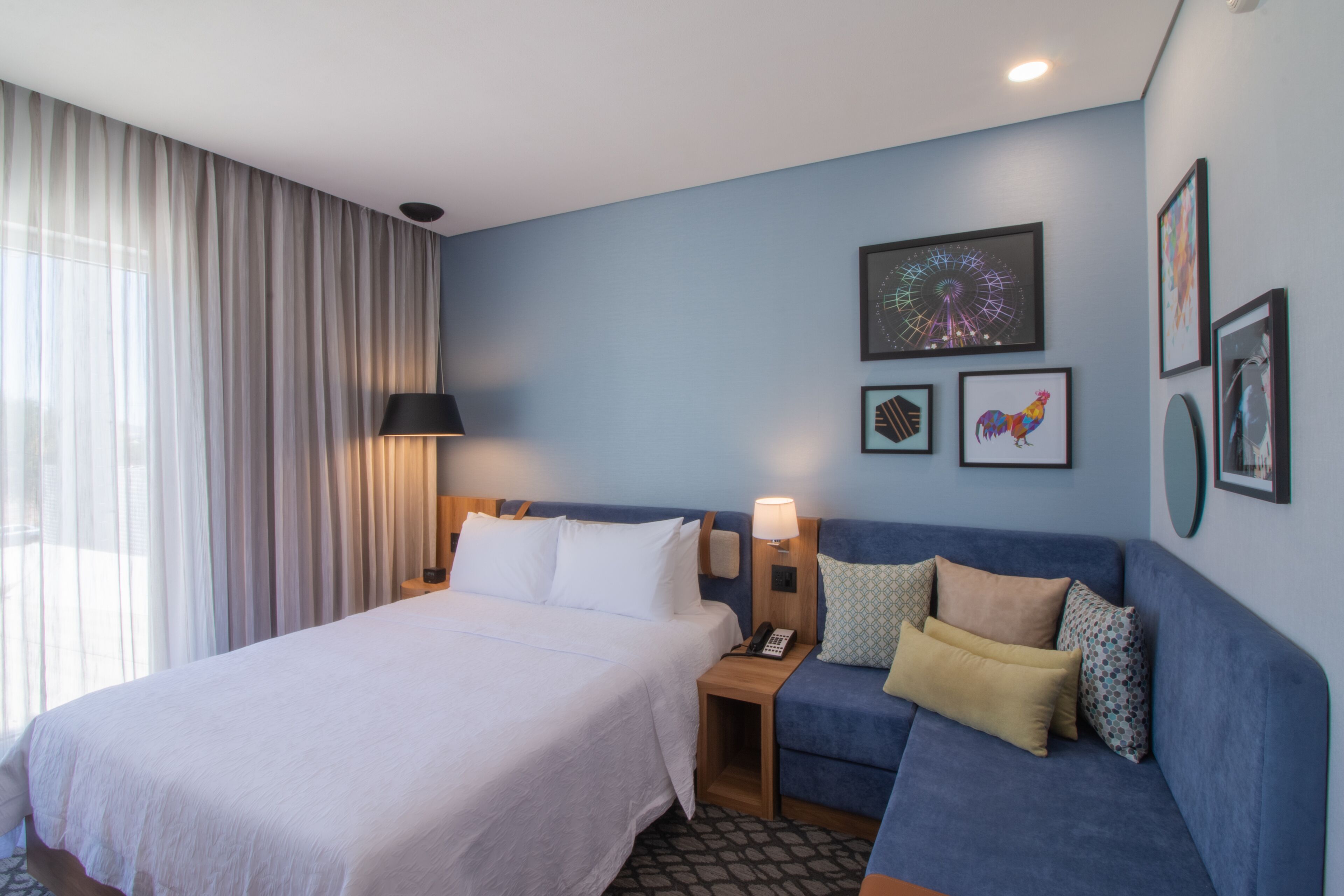 A hotel room with a bed, a blue sofa, a nightstand with a lamp, and framed pictures on the wall.