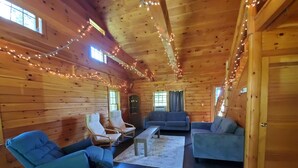 A cozy cabin interior with wooden walls, a sofa, two armchairs, a coffee table, and string lights. - Greenwood At Entangled Acres - Log Cabin w\WiFi on 16 Acre Animal Sanctuary (Owego)