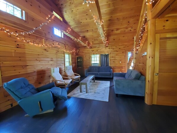 Un interior de cabaña acogedor, con paredes de madera, un sofá, dos sillones, una mesita de centro y luces colgantes.